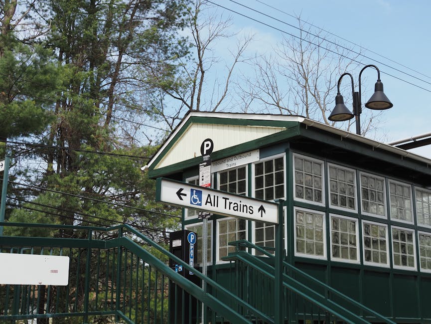 Exterior view of a small train station building with large glass block windows and a green and white wooden facade, situated amidst tall trees with leafless branches and overhead power lines. A stairc