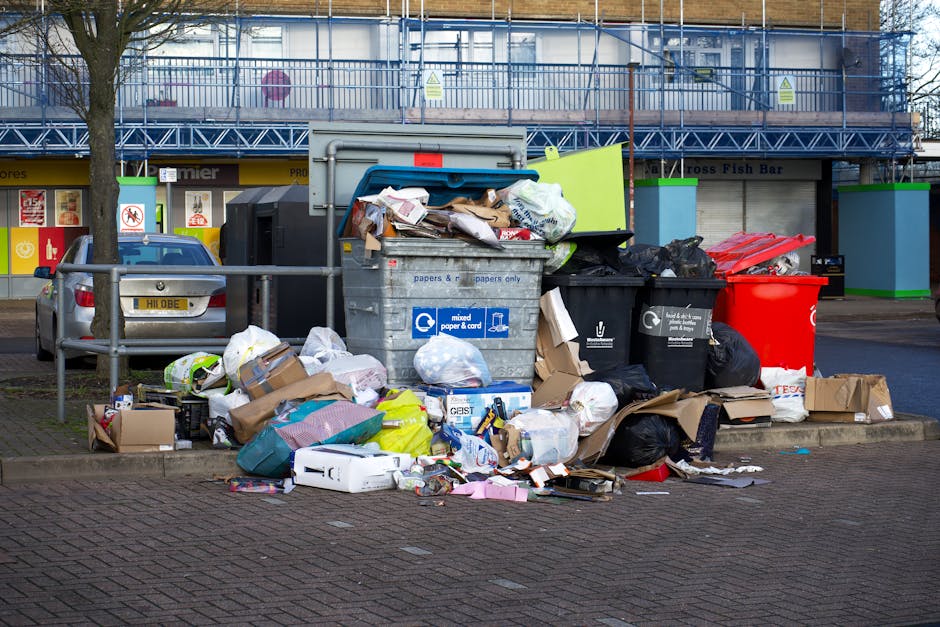 A collection of overflowing waste and recycling bins placed on a paved pavement area outside a commercial building, with various types of rubbish such as cardboard boxes, plastic bags, paper, and packaging materials spilling onto the ground. The bins include a large gray mixed paper and cardboard recycling container, a black bin for general waste, a black bin for recyclable materials, and a red bin, all positioned next to a metal railing. Behind the bins, a blue delivery van is being loaded or unloaded, with some furniture items or boxed goods possibly nearby. The building in the background features large signage, glass windows, and an external metal structure, with trees and parked cars visible in the surrounding area. The scene captures the process of waste disposal in an urban setting, relevant to moving and home relocation considerations handled by [COMPANY_NAME].