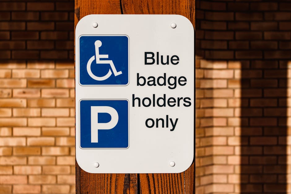 A rectangular parking sign mounted on a vertical wooden post, positioned outdoors against a backdrop of a brick wall. The sign features a blue square with a white wheelchair symbol indicating accessibility, and below it, a white background with bold black text that reads 'Blue badge holders only.' To the left of the text, there is a larger blue square with a white 'P' symbol, indicating parking. The sign's surface is clean and clearly visible in natural lighting, suggesting it is intended for designated parking areas reserved for blue badge holders, relevant to parking regulations for individuals with disabilities. This image supports content related to house removals and home relocation services, particularly highlighting access or parking considerations in the context of a professional moving process, such as those provided by Man with Van Ruxley, as seen in the website domain.