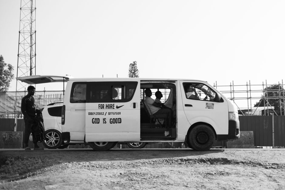 A black-and-white photograph of a white man with van Ruxley vehicle parked outdoors, with the sliding side door open revealing the interior and passengers seated inside. The van is positioned on a pav