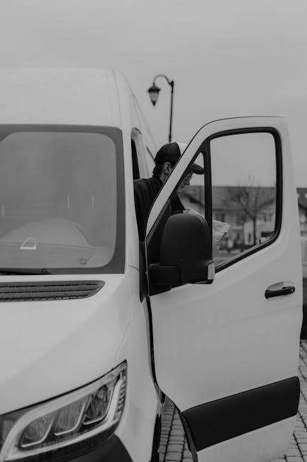 A black-and-white photograph of a white man with van Ruxley vehicle parked outdoors, with the sliding side door open revealing the interior and passengers seated inside. The van is positioned on a pav