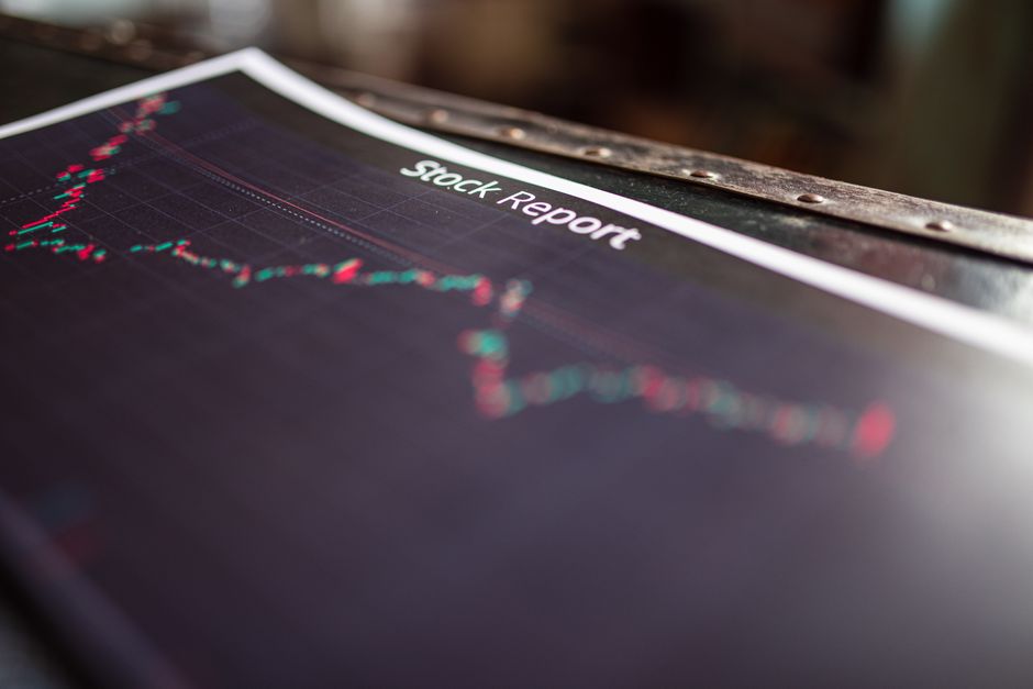 Close-up image of a financial stock report document placed on a wooden surface, showing a downward-trending line graph with red and green data points, printed on dark paper with a white header reading 'Stock Report'. The report is partially covered by a transparent plastic sheet, with a metal clip securing it at the top. The background includes a blurred view of a workspace, with natural light illuminating the scene, suggesting an environment where business or financial analysis may occur. This setting could be associated with planning or assessing business stock movements during home or office relocation preparations, relevant to professional removals and moving services like those offered by Man with Van Ruxley.