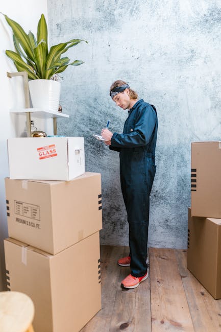 A person wearing a blue jumpsuit and protective goggles is standing indoors next to a stack of cardboard boxes, some labeled with descriptions such as 'glass.' The individual is holding a pen and appears to be taking inventory or noting details on a clipboard or notepad. The environment features a textured light-colored wall and wooden flooring, with a tall white shelf against the wall holding a large potted plant with broad green leaves. Additional boxes and packaging materials are visible nearby, indicating an organized packing or moving process. This scene captures elements of home relocation tasks, including packing, inventory management, and preparation for furniture transport, as part of the moving services offered by Man with Van Ruxley, supporting efficient and professional house removals.