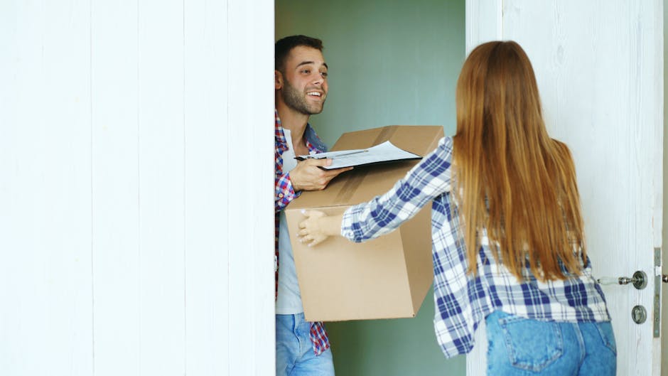 A man with dark curly hair and a beard, wearing a teal T-shirt with a logo on the upper left side, is standing indoors next to some cardboard moving boxes, including one labeled 'FRAGILE GLASS'. He is holding a clipboard and a pen, appearing to take notes or check items during the home relocation process. The room has a textured light blue wall, and in the background, there's a white shelf with a large potted plant with long, green leaves. The boxes are arranged on the floor, and some are stacked, suggesting packing for a house move. This scene illustrates the packing and moving stages involved in furniture transport and house removals, consistent with professional services like Man with Van Ruxley.
