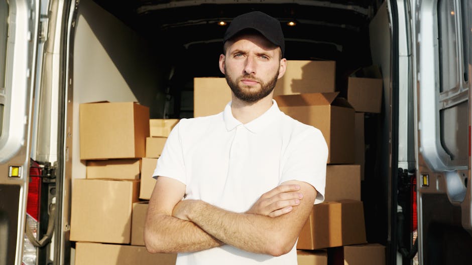 A man wearing a white t-shirt, light blue jeans, white sneakers, and a black cap is in the process of loading or unloading medium-sized cardboard boxes onto a white commercial van parked on the street