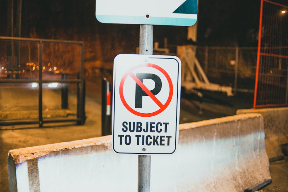 A close-up view of a white rectangular parking restriction sign with a bold black letter 'P' crossed out by a red circle and slash symbol, mounted on a metal pole. Below the symbol, the sign reads 'SUBJECT TO TICKET' in black capital letters. In the background, there is a concrete barrier and an orange safety fence, indicating a restricted or construction area, with dim street lighting illuminating the scene at night. This setting likely relates to road closure or parking permit regulations that could impact house removals or furniture transport, as managed by Man with Van Ruxley. The environment suggests a controlled zone for relocating items or vehicles, aligning with local permit rules for home relocation logistics.