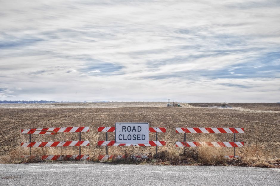 A rural scene featuring a road closure barrier with red and white striped metal panels and a central white sign displaying 'ROAD CLOSED' in black letters. The barrier is positioned at the edge of a paved road, leading into an open, flat field with dry, brown grass and no visible structures or vehicles nearby. In the background, a distant landscape with a few small buildings or construction equipment can be seen under a partly cloudy sky. The setting suggests an area undergoing road work or temporary access restriction, relevant to logistics and moving operations by Man with Van Ruxley, especially when coordinating house removals and furniture transport in areas with road closure or permit rules.