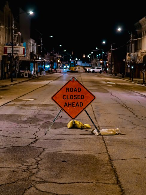 A close-up view of a white rectangular parking restriction sign with a bold black letter 'P' crossed out by a red circle and slash symbol, mounted on a metal pole. Below the symbol, the sign reads 'SUBJECT TO TICKET' in black capital letters. In the background, there is a concrete barrier and an orange safety fence, indicating a restricted or construction area, with dim street lighting illuminating the scene at night. This setting likely relates to road closure or parking permit regulations that could impact house removals or furniture transport, as managed by Man with Van Ruxley. The environment suggests a controlled zone for relocating items or vehicles, aligning with local permit rules for home relocation logistics.