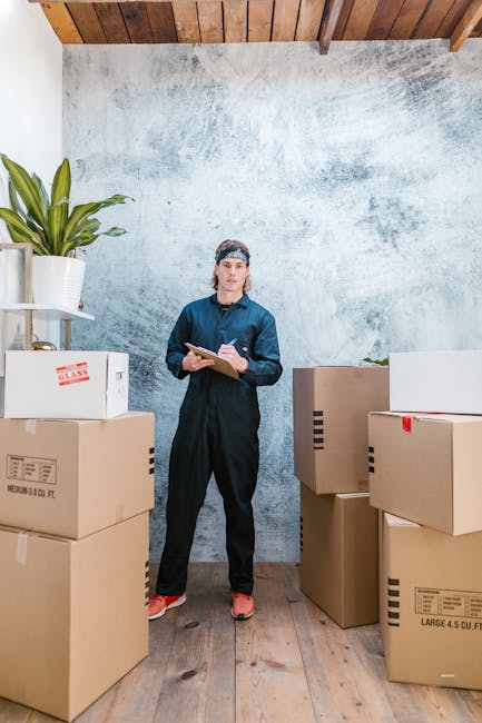 A young woman wearing a navy blue jumpsuit and a headband stands inside a room with a textured light grey wall and wooden ceiling beams. She holds a clipboard and pen, appearing to review or manage packing and moving arrangements. Around her are several large cardboard boxes, some labeled with capacity and contents, arranged on a wooden floor. In the background, there's a white shelf with a potted plant, indicating an indoor area prepared for house relocation. The scene is well-lit with natural or soft artificial lighting, suggesting an organized packing process. This setup reflects a professional environment for coordinating furniture transport and packing for a home move, which aligns with the services provided by Man with Van Ruxley as highlighted on their Ruxley estate packing and parking guide page.