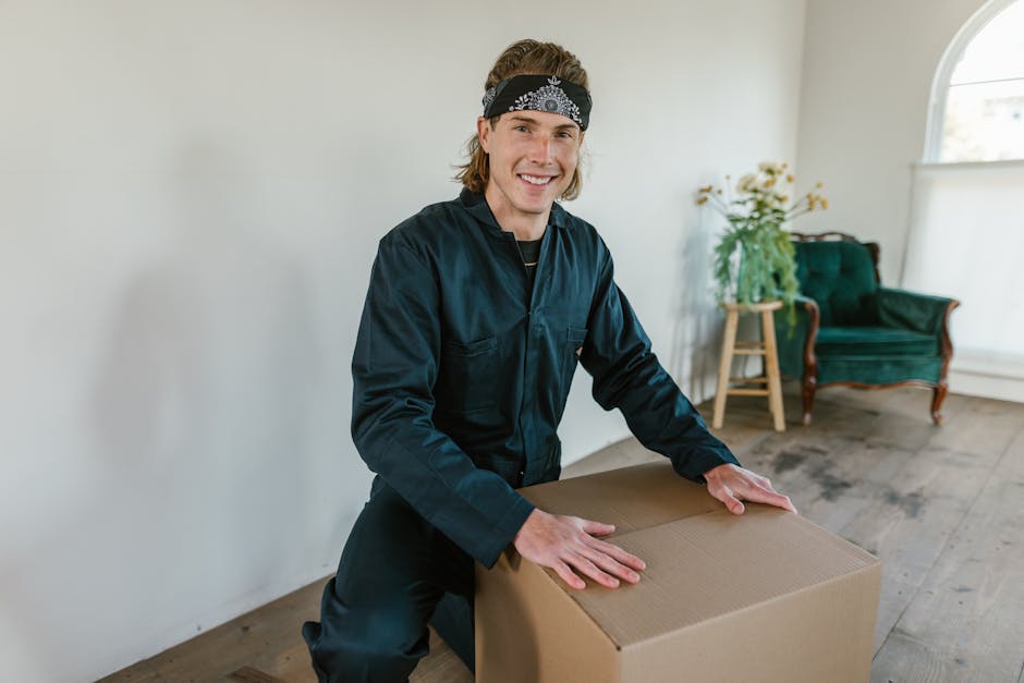 A male mover with shoulder-length blonde hair, wearing a black work uniform and a black bandana with white patterns, is smiling while lifting a medium-sized cardboard box during a home relocation process inside a well-lit room. The room features white walls, wooden flooring, and natural light coming from a large arched window. In the background, there is a green velvet armchair with wooden legs, a wooden side table holding a potted plant with white flowers and green foliage, and a dark-colored rug partially visible on the floor. The mover’s posture indicates he is carefully handling the box, which is sealed with packing tape. This image represents professional packing and furniture transport activities performed by Man with Van Ruxley, as part of their house removals services, illustrating the loading process of household items for an organized move.
