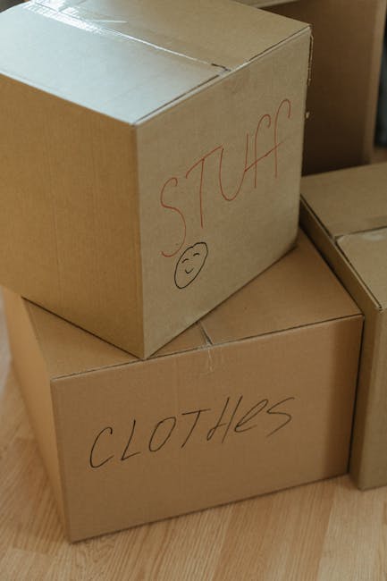 Close-up image showing three stacked cardboard moving boxes in a home environment, with the top box labeled 'STUFF' in red marker alongside a smiley face, and the bottom box labeled 'CLOTHES' also in red marker. The boxes are placed on a wooden floor, and the surrounding area appears to be part of a room prepared for moving or packing. The boxes are sealed with packing tape, indicating they are packed and ready for transport as part of a home relocation or furniture transport process, with the branding of Man with Van Ruxley associated with professional removals services.