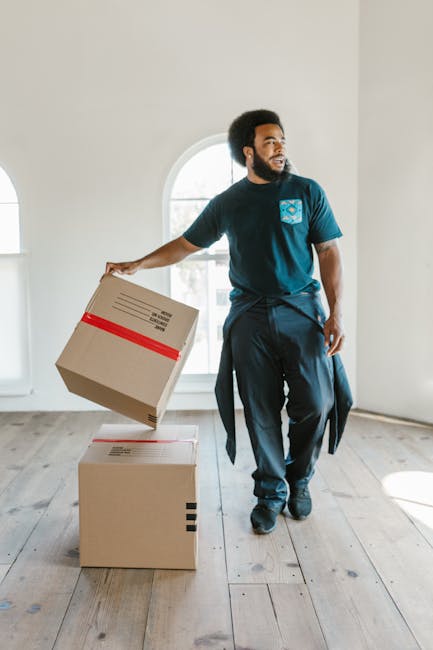 A man with dark curly hair and a beard, dressed in a navy blue t-shirt and matching trousers, is inside a bright, spacious room with white walls and a large arched window allowing natural light. He is holding a medium-sized cardboard box wrapped with red packing tape in his right hand, preparing to move or load it. On the floor in front of him, there is a larger cardboard box, also sealed with red tape, positioned on a light wooden floor. The scene captures the process of packing or furniture transport related to home relocation, with the man appearing to be in the midst of packing or moving supplies, consistent with professional removals services provided by Man with Van Ruxley. The room is empty apart from these boxes, emphasizing the focus on packing and transport logistics typical in house removals and furniture transport tasks.