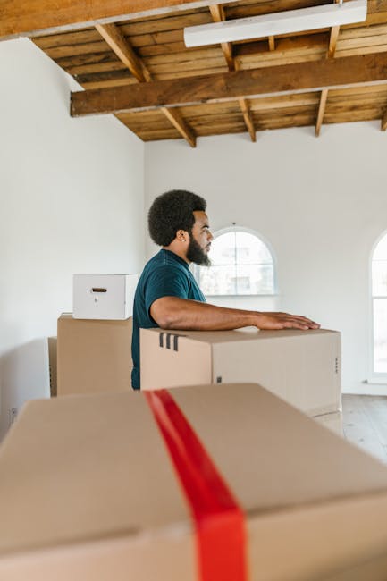 A man with dark curly hair, beard, and wearing a teal T-shirt stands inside a room with white walls and a wooden ceiling. He is positioned next to a stack of plain cardboard moving boxes, one of which he is resting his hands on. The boxes vary in size, with some featuring cut-out handles. The room has natural light coming through arched windows, revealing a residential setting. The scene depicts a home relocation process, with the man preparing for furniture transport by organizing and handling boxes. Visible in the background are ceiling beams and an unfinished wooden structure, indicating an interior space that may be undergoing renovations or pre-move packing. The environment appears clean and orderly, suitable for professional removals by [COMPANY_NAME], supporting efficient packing and loading during a same-day move.