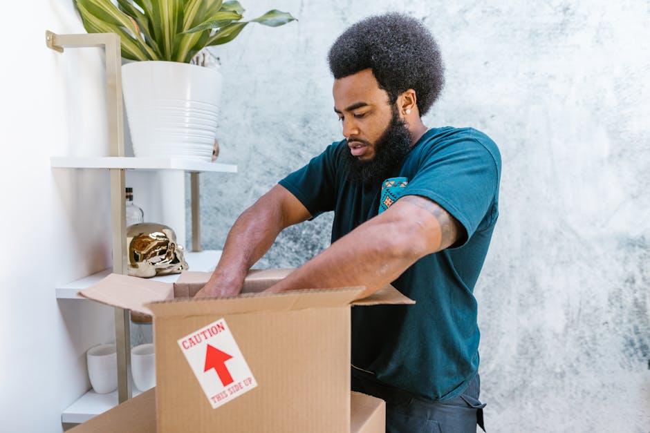 Two movers from Man with Van Ruxley are seen inside a well-lit room with large arched windows, engaged in a home relocation process. One mover, with curly hair and a beard, is holding a large cardboard box with red packing tape, standing near three other boxes stacked on the wooden floor. The second mover, wearing a headband and dark clothing, is speaking and holding a medium-sized cardboard box. The room contains additional boxes, some placed on the floor near the movers, indicating an ongoing packing and moving operation. The scene shows elements of furniture and packing materials, with soft natural light entering through the windows, and a vintage armchair visible at the edge of the room. The environment suggests a professional furniture transport or complete home packing process as part of a removal service.