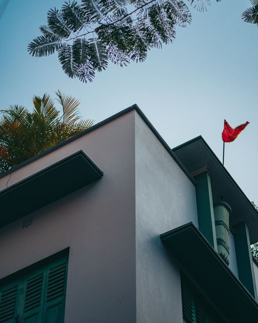 A low-angle view of a modern residential building with white exterior walls and teal window shutters, captured during daylight with clear blue sky overhead. The building has a flat roof, and a red flag is mounted on a pole attached to the side of the structure. Surrounding the building, there are large green tropical leaves and palm fronds partially visible at the top of the image, indicating an outdoor setting. The photograph emphasizes the clean lines and contemporary design of the property, which is in the process of being prepared for home relocation or furniture transport, as part of professional removals services by Man with Van Ruxley. Inside the property, visible through the open window shutters, are packing materials and furniture, while outside, a moving vehicle or loading area is not directly visible but implied by the context of loading and transportation. The scene reflects the initial stages of a house move, including structural features and the outdoor environment relevant to loading and logistics.